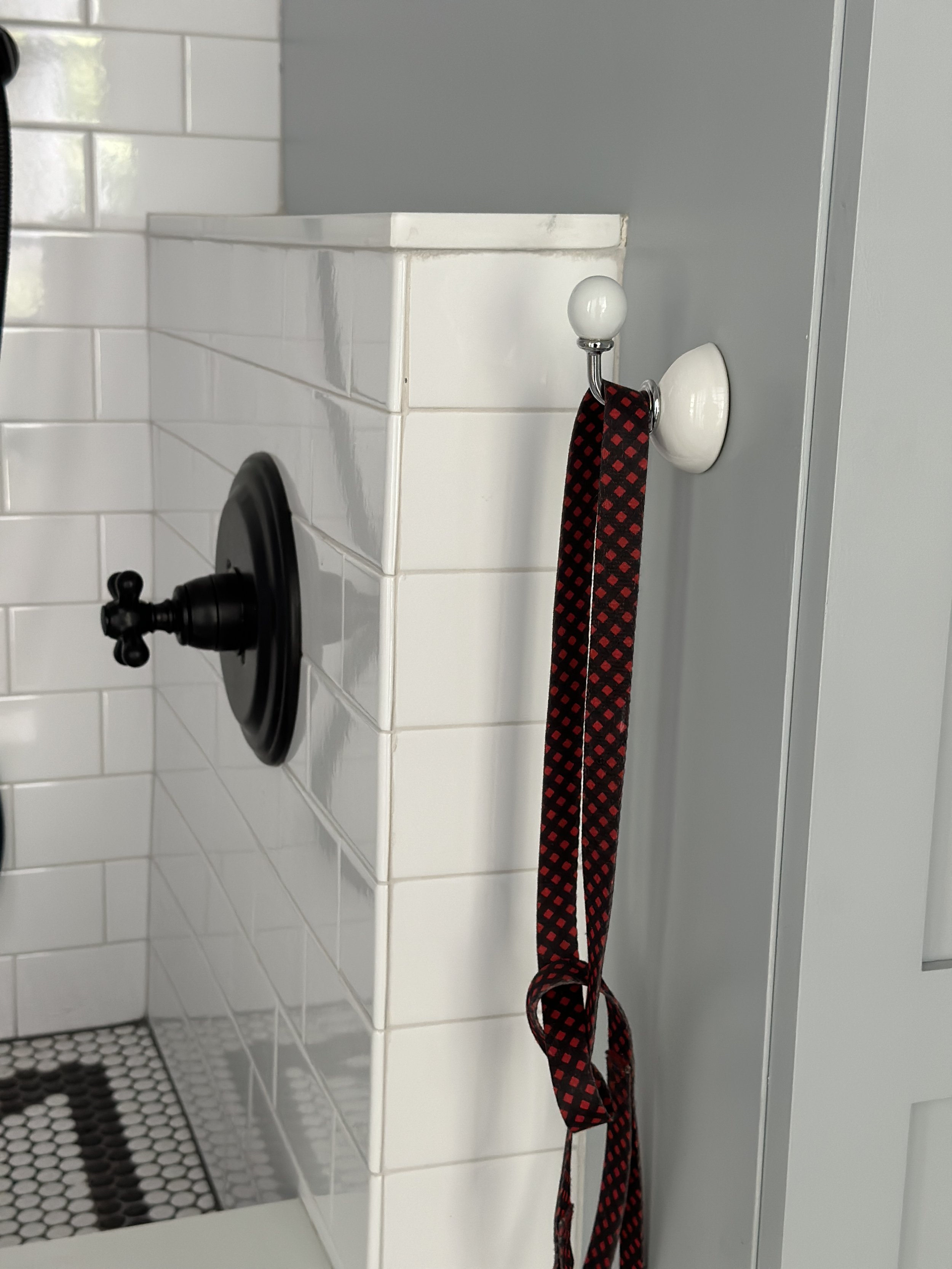 Red checked dog leash hanging on a wall hook beside the white-tiled dog shower, with a black handheld sprayer and a hexagon tile floor, in a Westfield, New Jersey mudroom by Ellie Mroz Design.