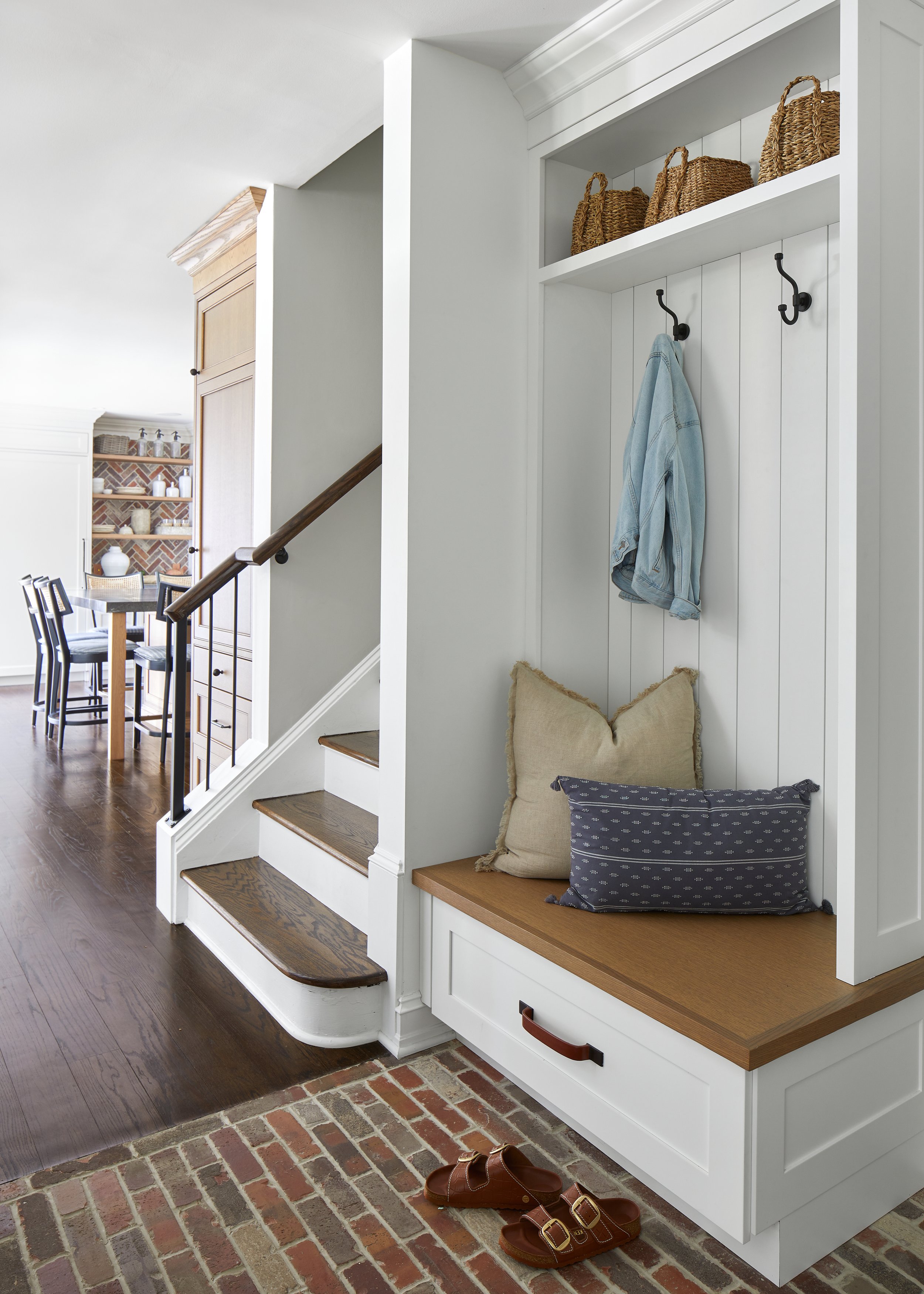 Built-in mudroom bench with a white oak seat, brass hooks, woven baskets on the shelf above and a brick floor, beside the stair in a 1929 Westfield, New Jersey home renovated by Ellie Mroz Design.