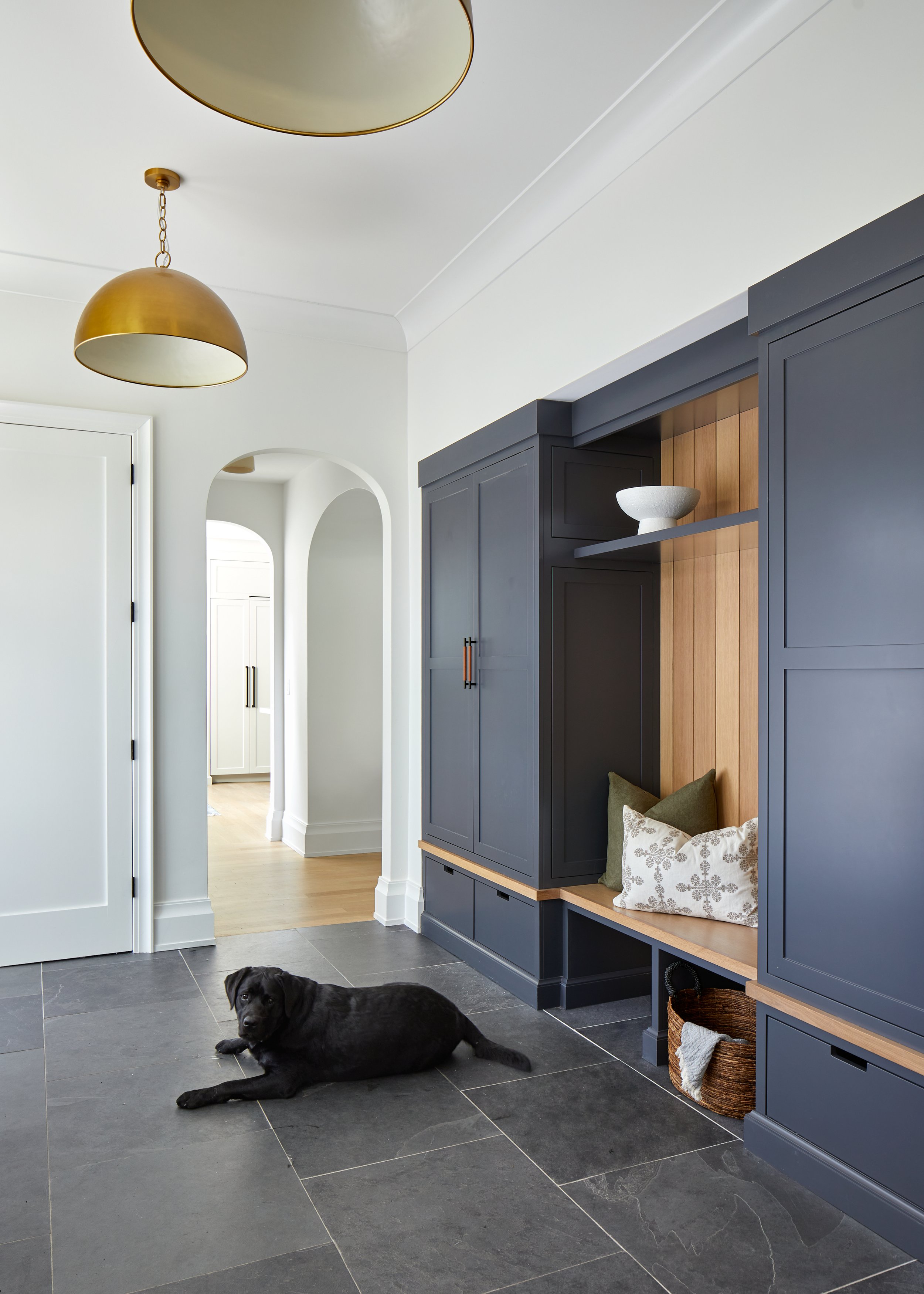Mudroom with navy built-in lockers, a white oak bench with baskets below, brass dome pendants and slate flooring, with a black Labrador lying by the arched doorway, in a Westfield, New Jersey renovation by Ellie Mroz Design.