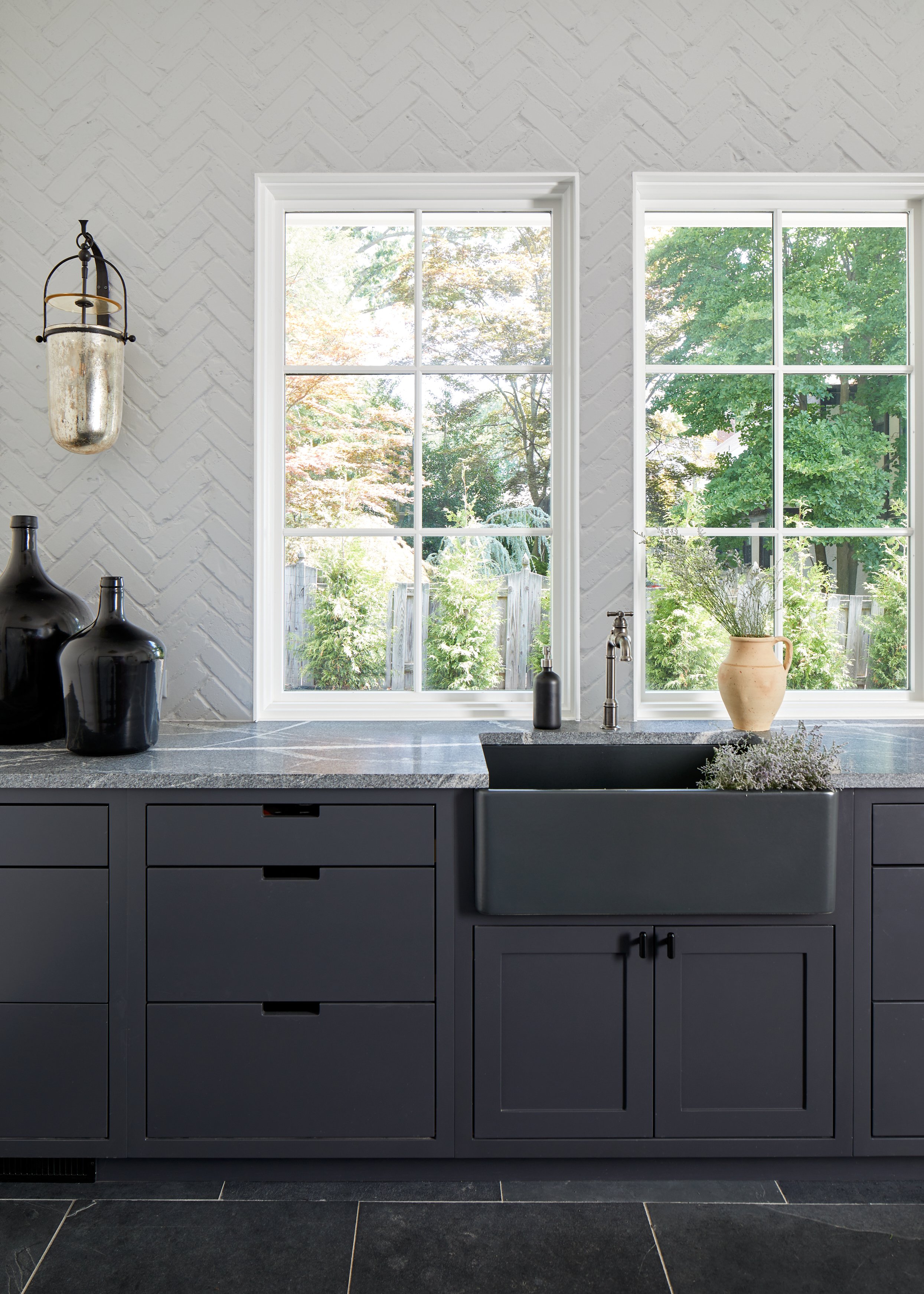 Mudroom sink wall with navy cabinetry, a black apron-front sink, white herringbone tile, a glass lantern pendant and slate floors in a Westfield, New Jersey renovation by Ellie Mroz Design.