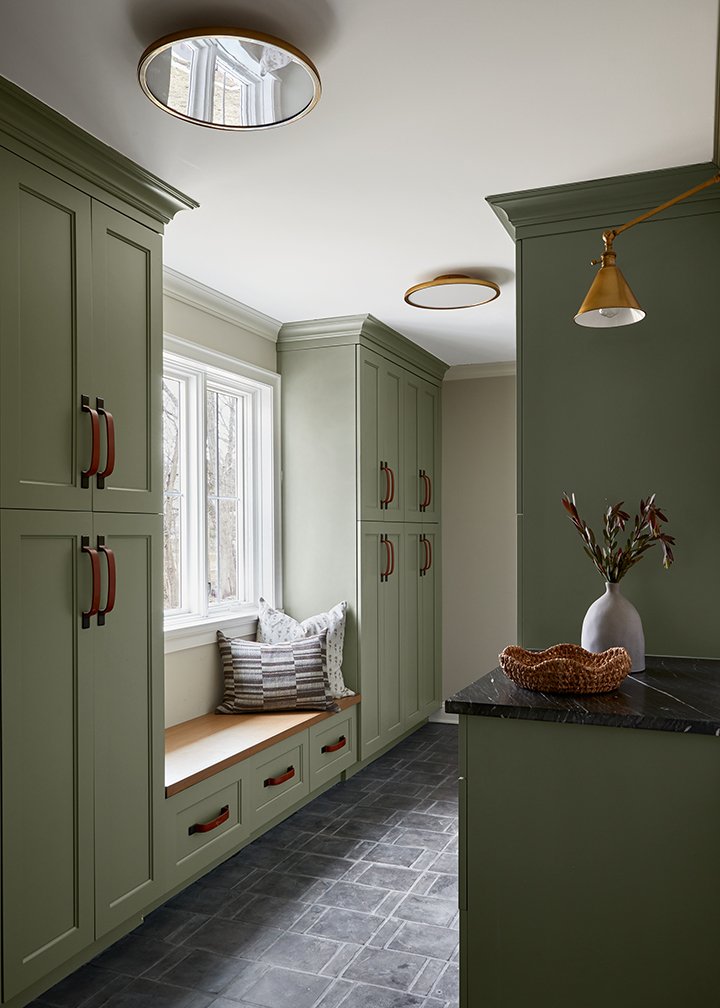 Olive green mudroom lockers and a built-in bench with a wood seat, brass sconces and a round mirror above slate floors in a Westfield, New Jersey renovation by Ellie Mroz Design.