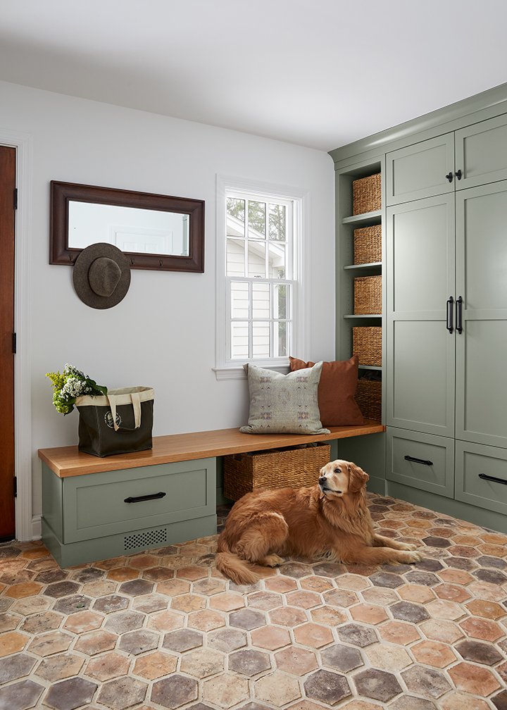 Sage green mudroom cabinetry with a wood bench, woven baskets and a terracotta hexagon tile floor, with a golden retriever lying in front of the bench, in a Westfield, New Jersey renovation by Ellie Mroz Design.
