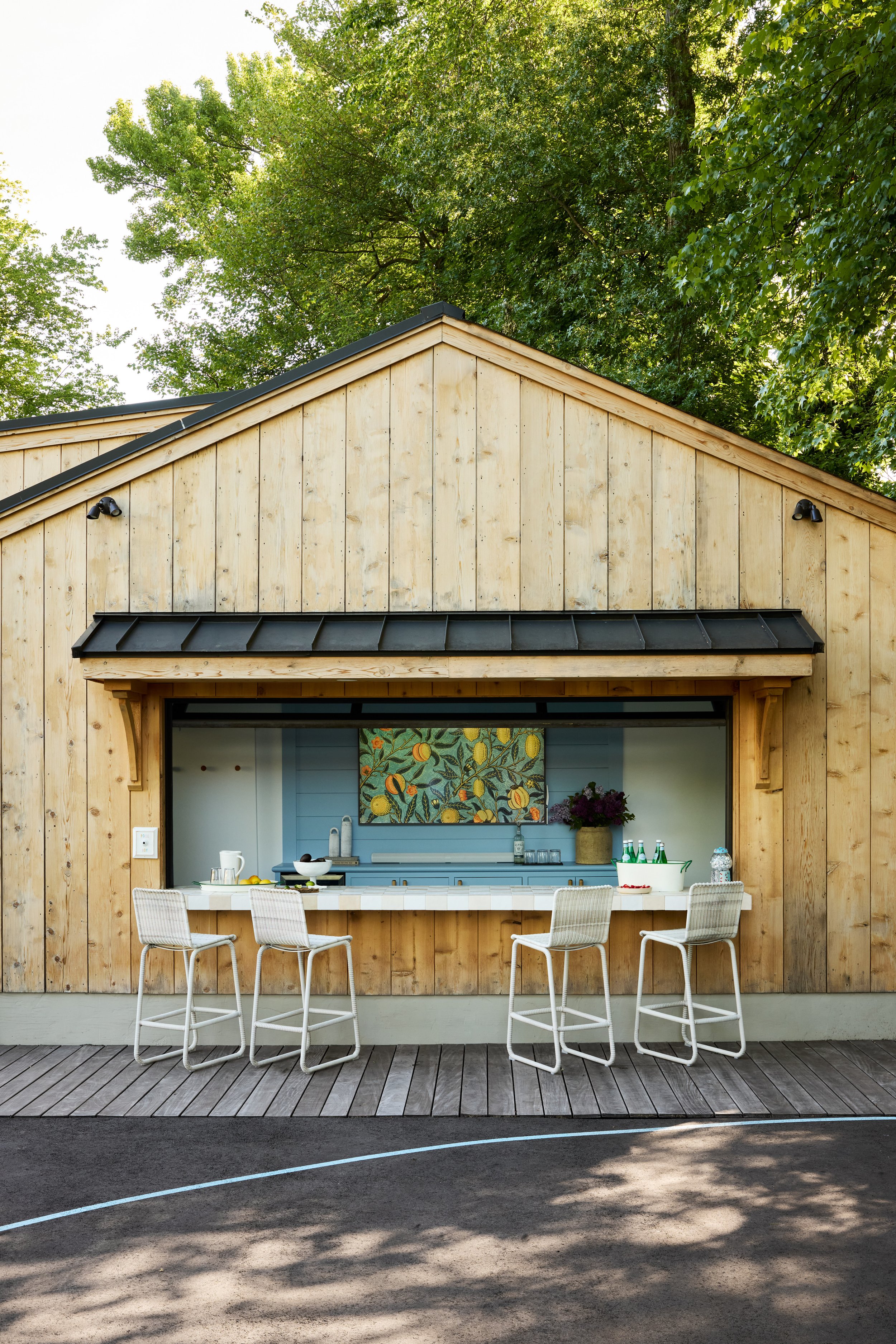Outdoor bar built into a cedar-clad pool house in Westfield, NJ, with blue cabinetry, a patterned tile counter and four wicker barstools by Ellie Mroz Design.